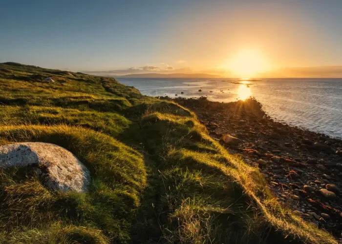 Sunset over a grassy coastline with rocks scattered along the shore and sunlight reflecting on the calm sea, capturing the tranquil beauty of the Summer Solstice Ireland on the longest day of the year.