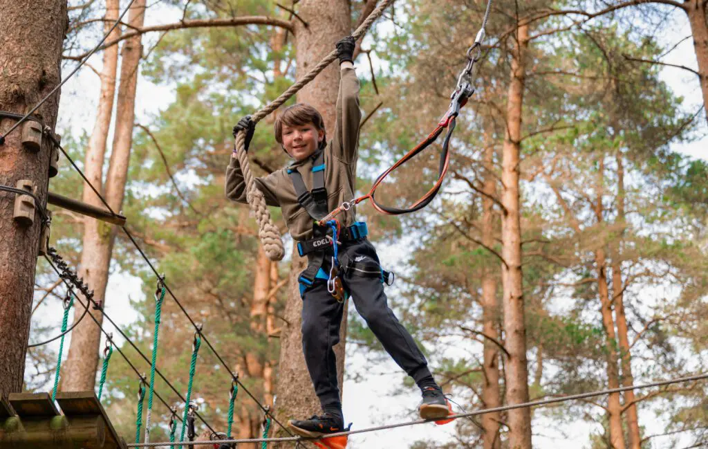 A boy with a safety harness crosses a rope bridge at Zipit adventure park, holding onto a rope above his head—a perfect summer activity for a fun family day out in the forest.