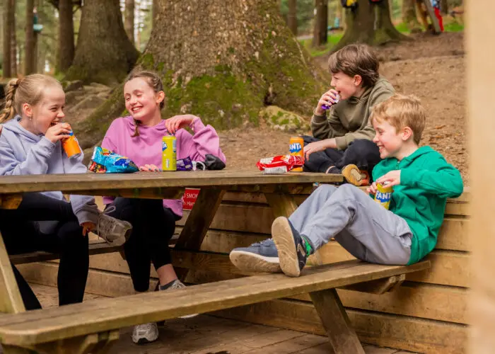 Four children sit at a wooden picnic table in a forest, laughing and drinking bottled sodas with snacks on the table—an ideal Summer Activity for a Family Day Out after an adventure at Zipit.