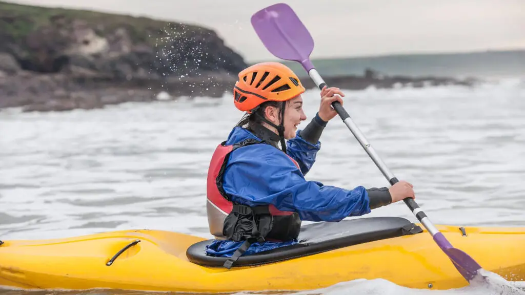 Person wearing a helmet and life jacket paddles a yellow kayak in choppy water near a rocky shoreline, embracing Irish family adventures and inviting everyone to get into nature.