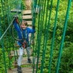 A child wearing a blue jacket and harness beams with joy while crossing a rope bridge, enjoying mid-term activities in the lush Lough Key Forest and Activity Park.