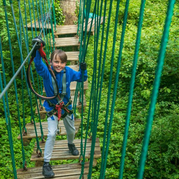 A child wearing a blue jacket and harness beams with joy while crossing a rope bridge, enjoying mid-term activities in the lush Lough Key Forest and Activity Park.