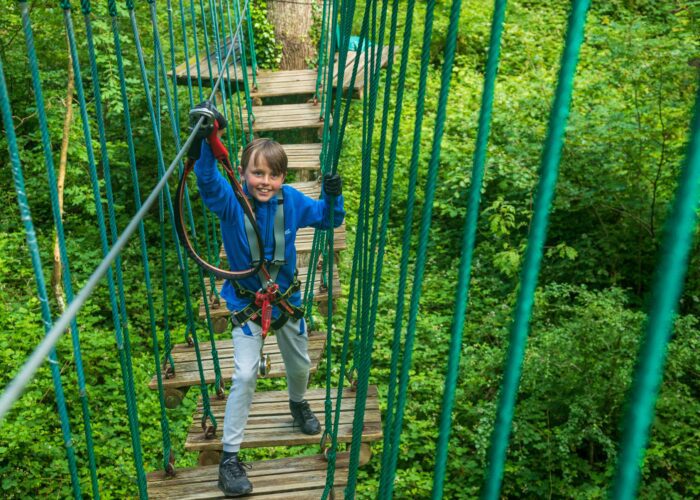 A child wearing a blue jacket and harness beams with joy while crossing a rope bridge, enjoying mid-term activities in the lush Lough Key Forest and Activity Park.