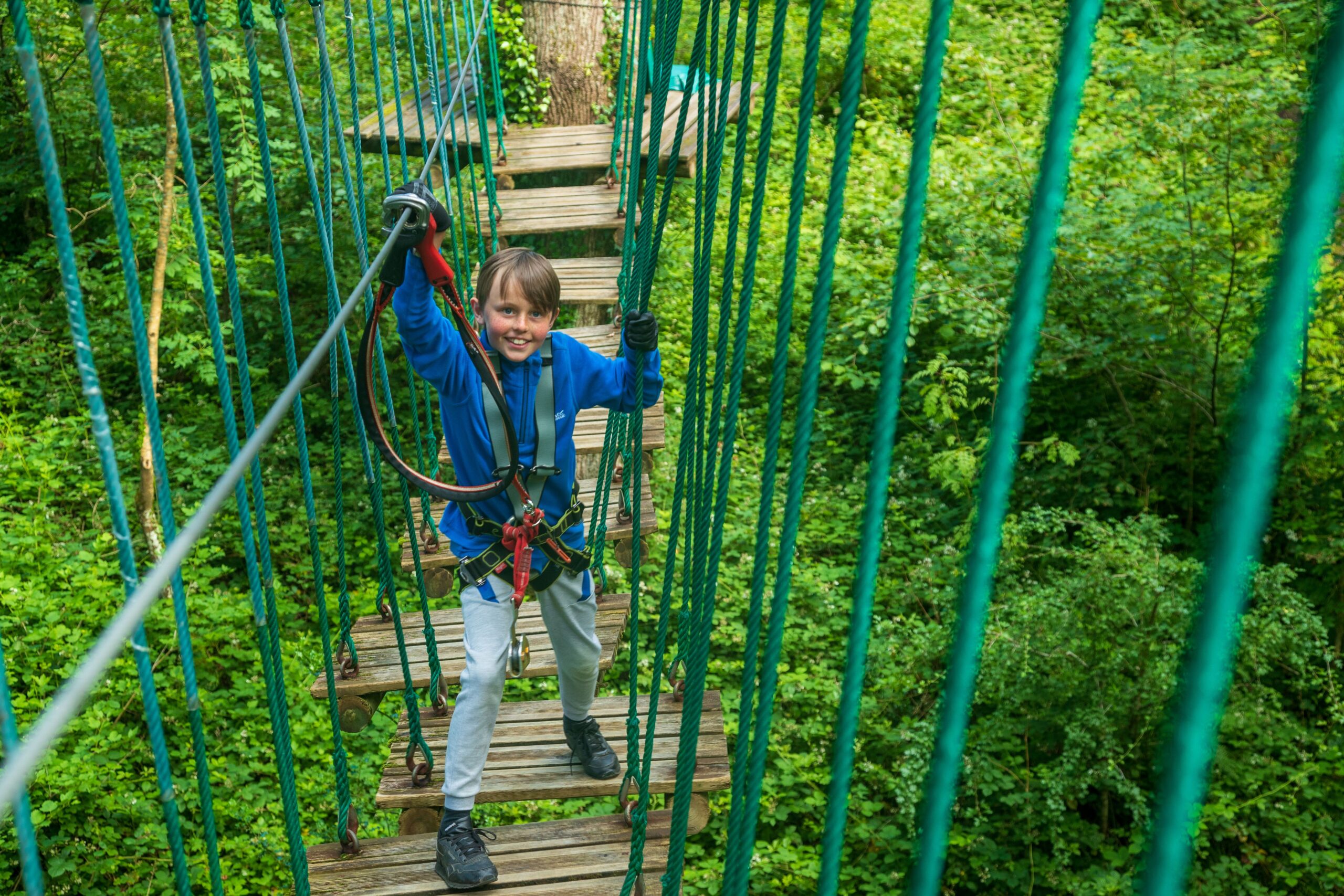 A child wearing a blue jacket and harness beams with joy while crossing a rope bridge, enjoying mid-term activities in the lush Lough Key Forest and Activity Park.