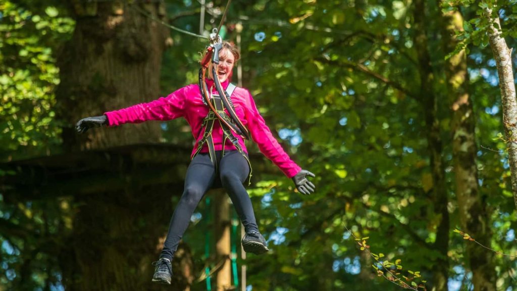 A person wearing a pink jacket and black gloves smiles while zip-lining through a forest.
