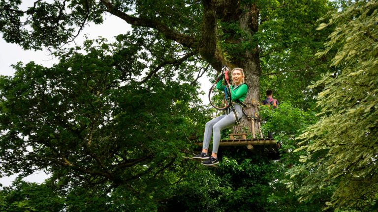 A person in green clothing zip-lining through trees with a wooden platform in the background. A person in green clothing zip-lining through trees with a wooden platform in the background.