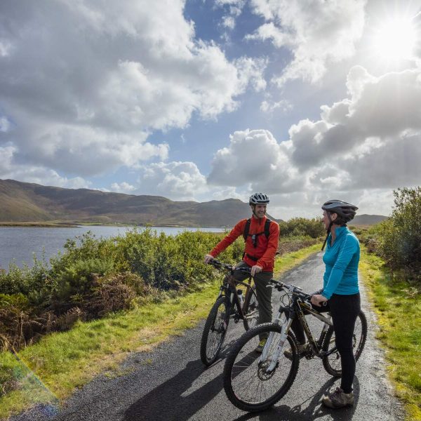 Two cyclists wearing helmets pause on a scenic lakeside road surrounded by hills and clouds, with sunlight breaking through. It's the perfect Bank Holiday Weekend escape, embracing the Great Outdoors.
