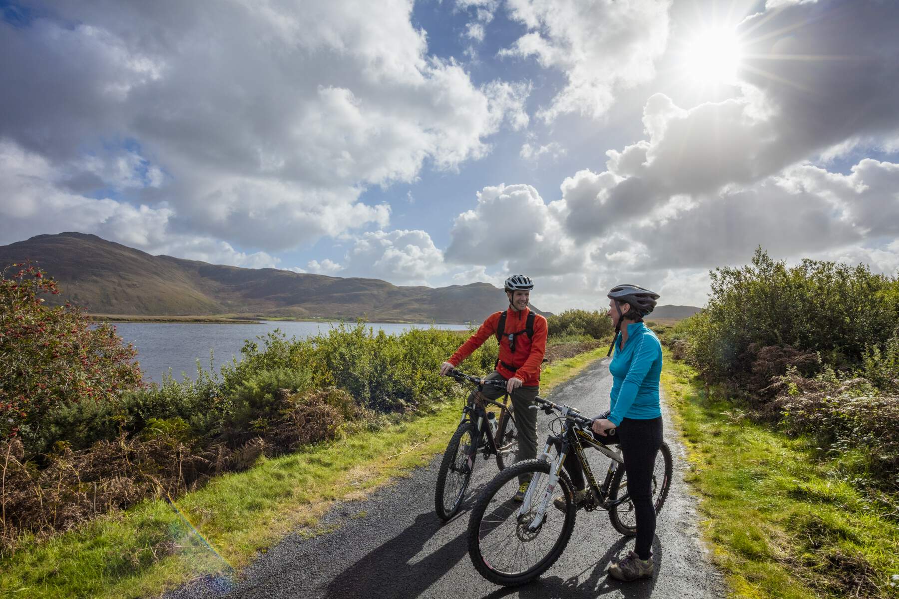 Two cyclists wearing helmets pause on a scenic lakeside road surrounded by hills and clouds, with sunlight breaking through. It's the perfect Bank Holiday Weekend escape, embracing the Great Outdoors.