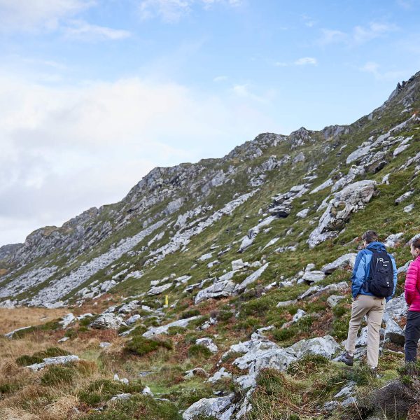 Three hikers explore the Great Outdoors, walking along a rocky, grassy trail between two hills under a partly cloudy sky. Perfect for a Bank Holiday Weekend adventure.