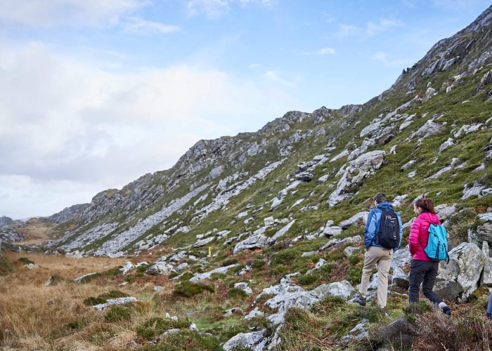 Three hikers explore the Great Outdoors, walking along a rocky, grassy trail between two hills under a partly cloudy sky. Perfect for a Bank Holiday Weekend adventure.