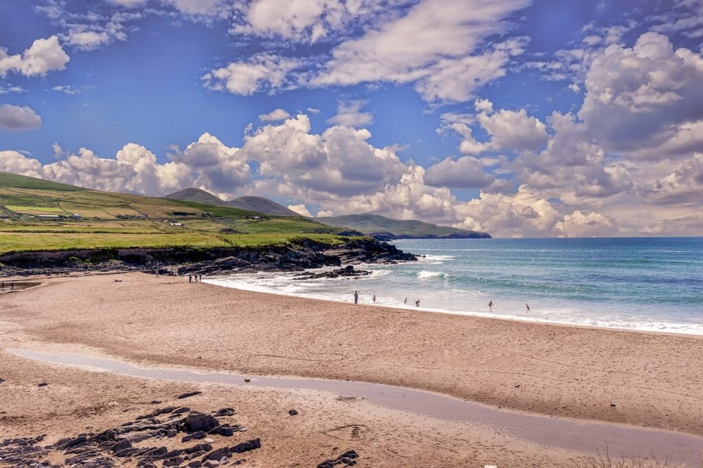 A sandy beach curves along the shoreline with waves, distant hills, grassy fields, and a partly cloudy sky. Several families are visible near the water, enjoying an end-of-summer adventure by the sea.