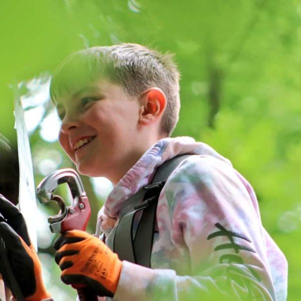Child in a climbing harness grips a carabiner while ascending a tree, surrounded by greenery.