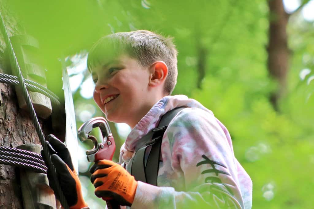 Child in a climbing harness grips a carabiner while ascending a tree, surrounded by greenery.
