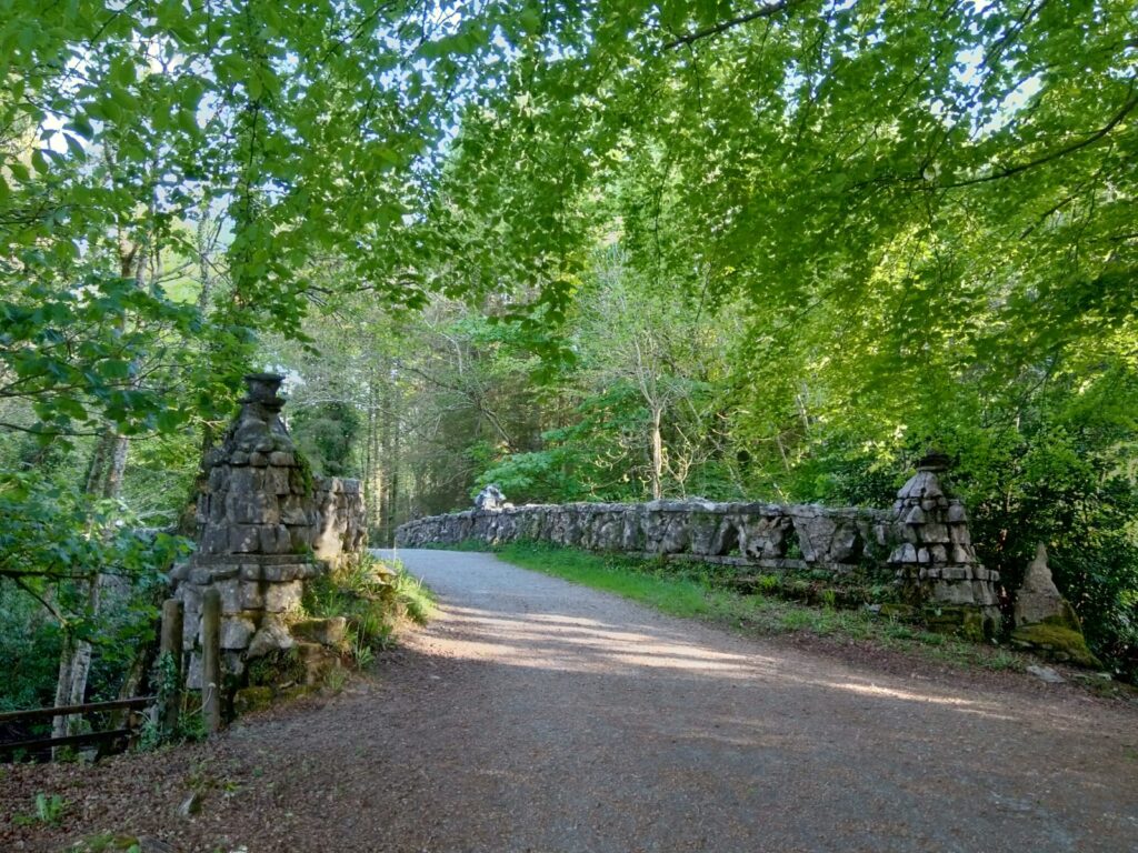 A stone bridge with rustic wooden railings crosses a dirt pathway surrounded by lush green trees, offering a gateway to beautiful forests in Roscommon, perfect for outdoor adventures.