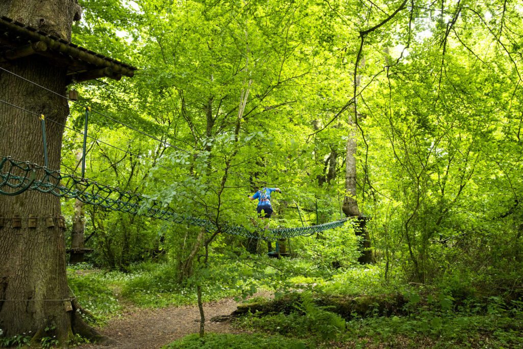 A person in a blue jacket crosses a rope bridge in the lush green canopy of Lough Key's adventure park, showcasing the thrilling beauty of the local area.