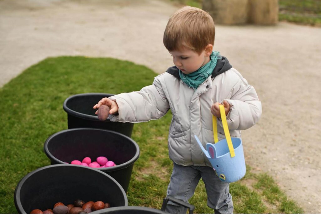 A child in a gray coat holds an egg while standing by buckets of colorful eggs, celebrating the Easter Egg-Stravaganza. He carries a blue basket with a mouse design, capturing the joy of Easter festivities.