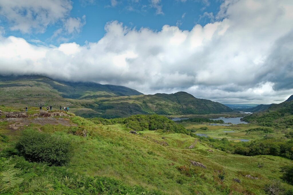 A lush green landscape in Ireland with rolling hills and a lake under a cloudy sky. Small groups of people are observing the scenery, savoring their Outdoor Escapes.