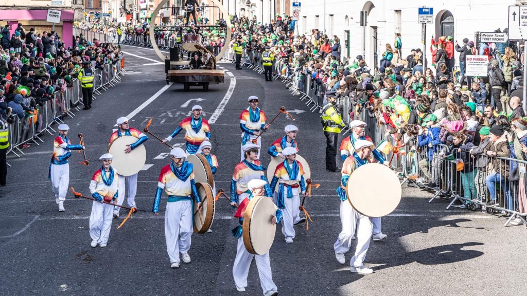 Performers in white costumes with drums march in a lively street parade, surrounded by crowds behind barricades on a sunny day.