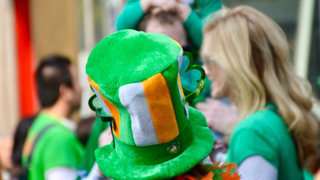 Person wearing a green top hat with orange and white stripes in a crowd, likely at a St. Patrick's Day event.