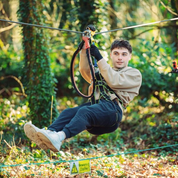 A person wearing casual clothes and safety gear glides along a zipline through a wooded area with a warning sign visible on the ground below.