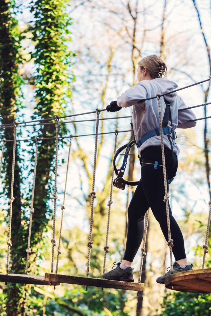 A person wearing a harness crosses a rope bridge in an outdoor adventure park surrounded by trees.