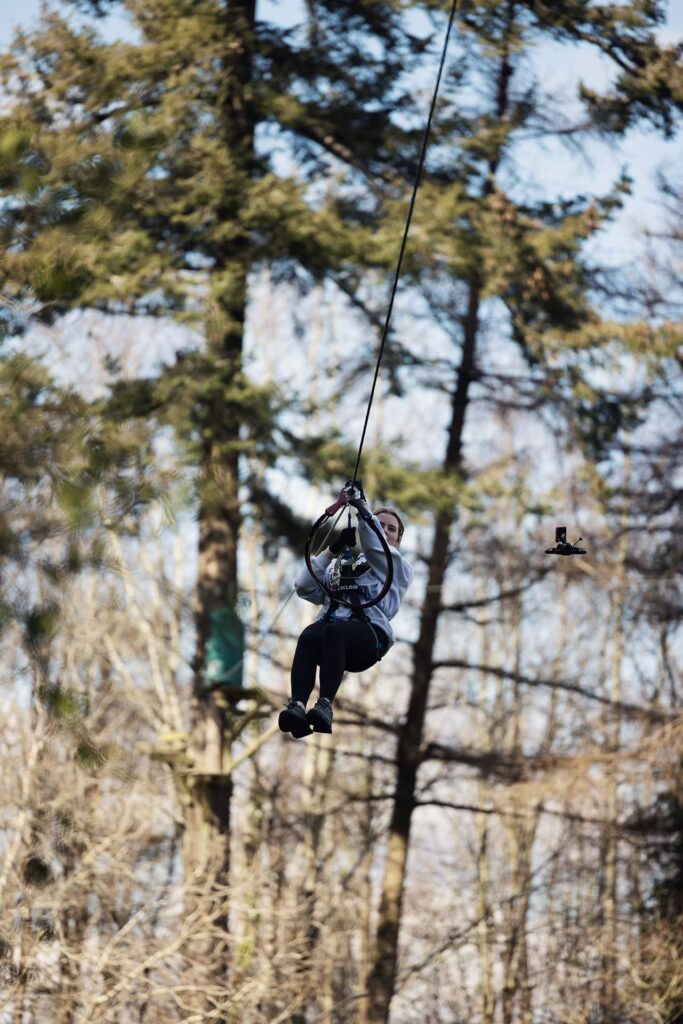Person riding a zip line through a forested area on a sunny day, with trees visible in the background.