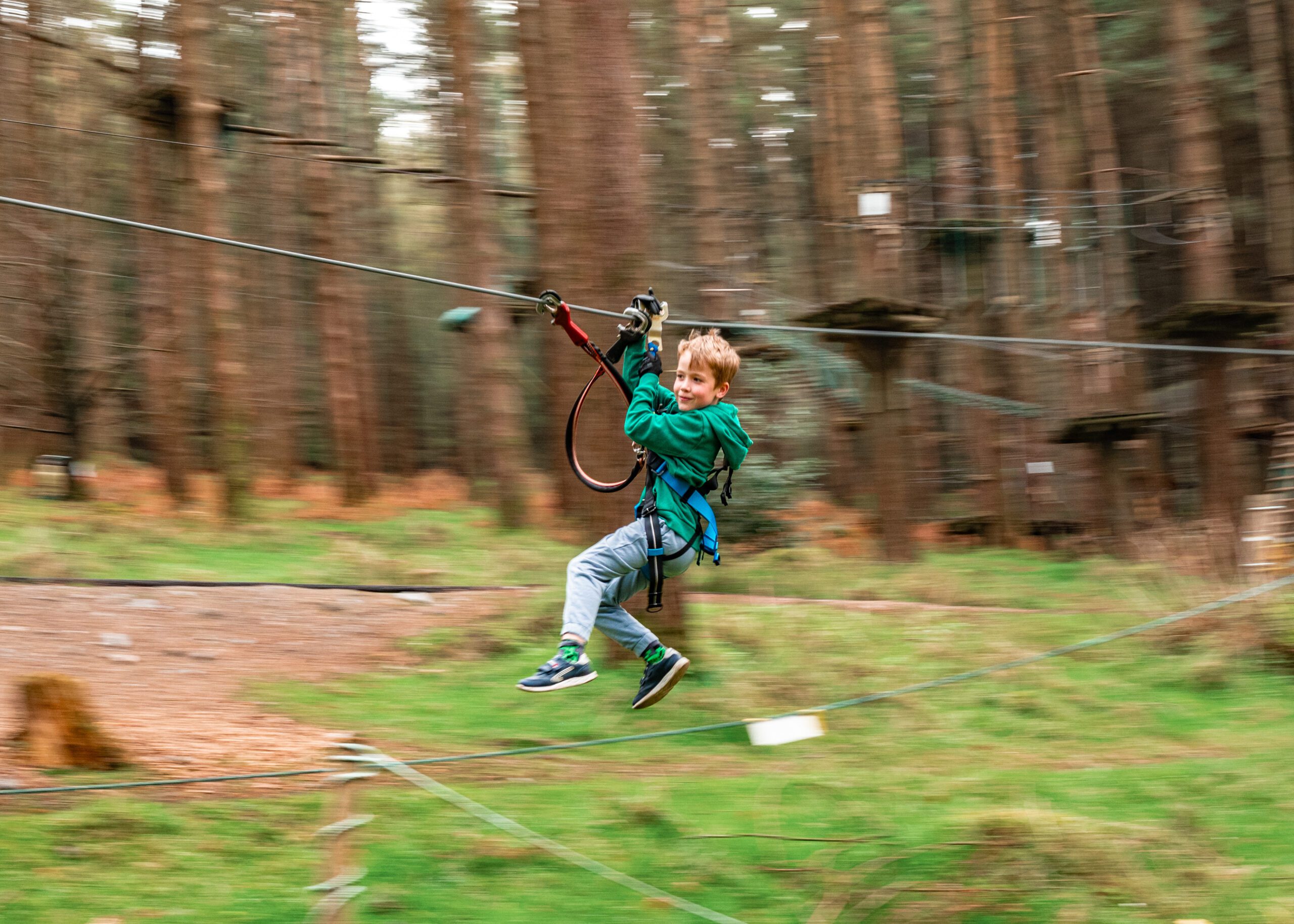 A child wearing a harness rides a zipline through a forested area, with trees and elevated platforms in the background.