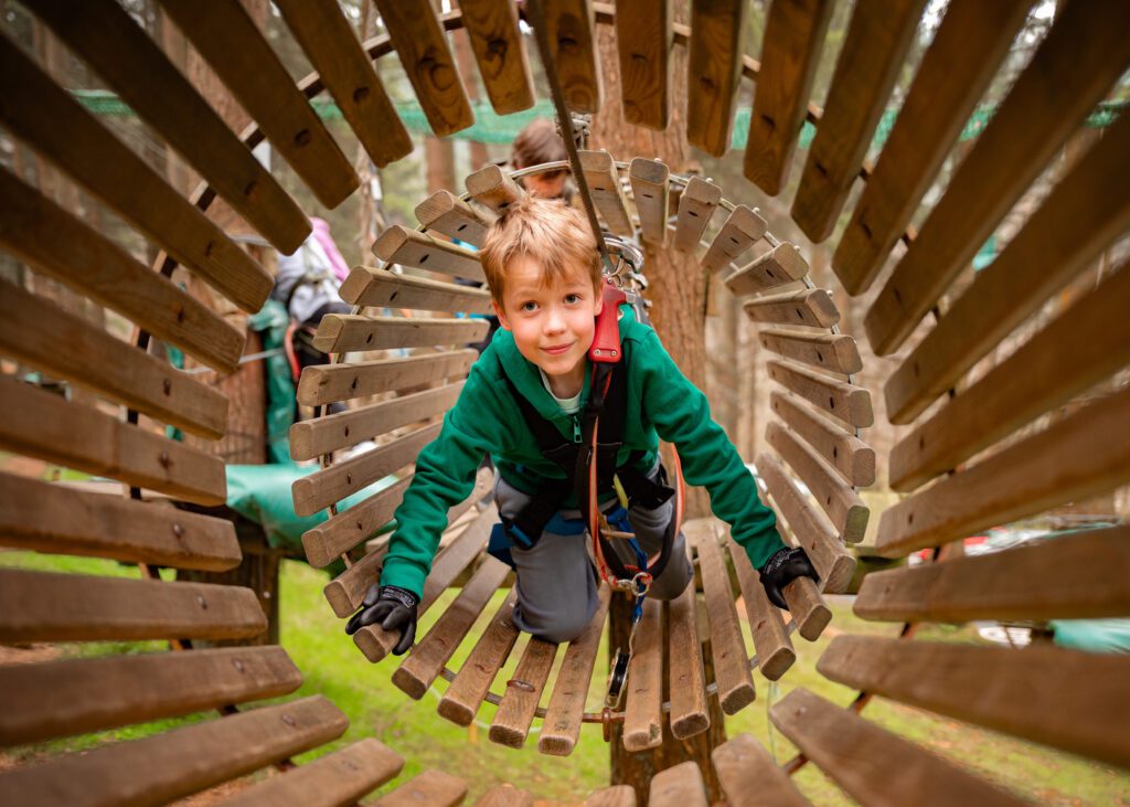 A child wearing a safety harness crawls through a wooden tunnel on an outdoor adventure course in a forest.