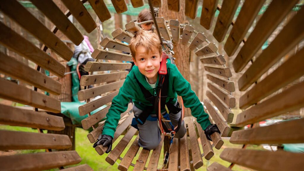 A child wearing a green jacket and safety harness crawls through a wooden tunnel structure in an outdoor adventure park.