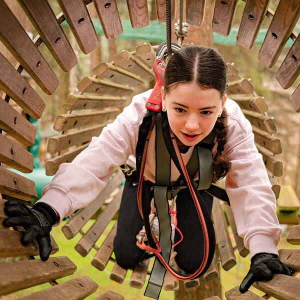 A young person with braided hair navigates a wooden tunnel obstacle while wearing safety gear and gloves at an outdoor adventure park.