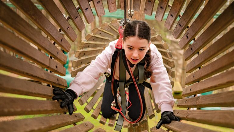 A young person with braided hair navigates a wooden tunnel obstacle while wearing safety gear and gloves at an outdoor adventure park. A young person with braided hair navigates a wooden tunnel obstacle while wearing safety gear and gloves at an outdoor adventure park.
