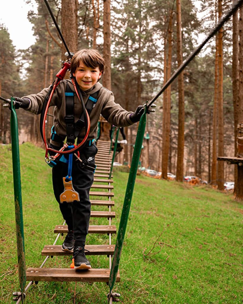 A child wearing safety gear crosses a suspended rope bridge in a forest adventure park.