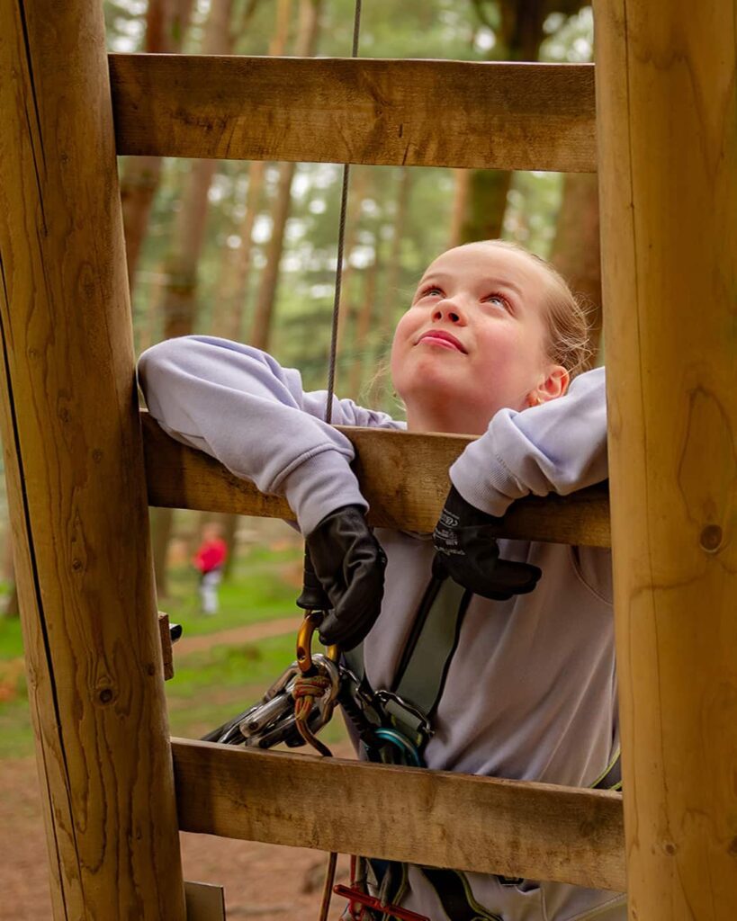 A child wearing a safety harness and gloves looks up while climbing a wooden outdoor structure in a forest.