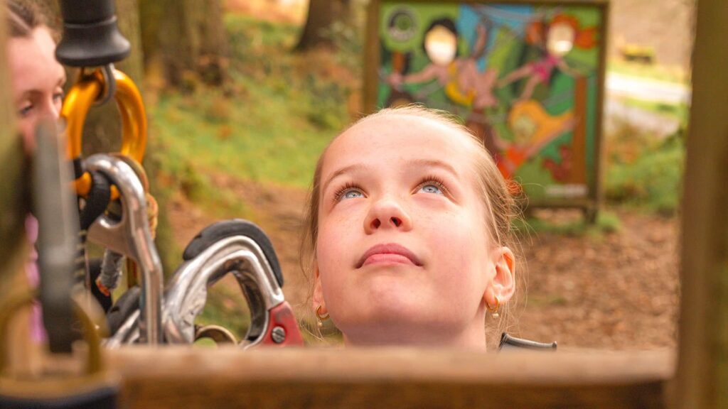 A young person looks upward outdoors near climbing equipment, with a colorful sign visible in the blurred background.
