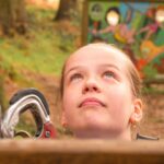 A young person looks upward outdoors near climbing equipment, with a colorful sign visible in the blurred background.