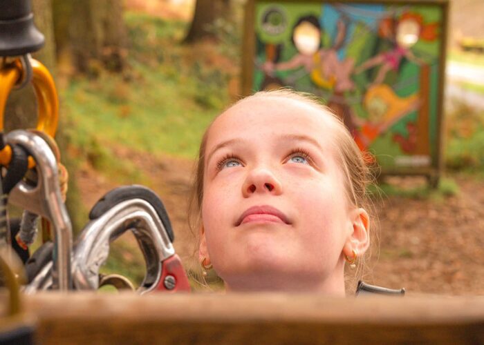 A young person looks upward outdoors near climbing equipment, with a colorful sign visible in the blurred background.