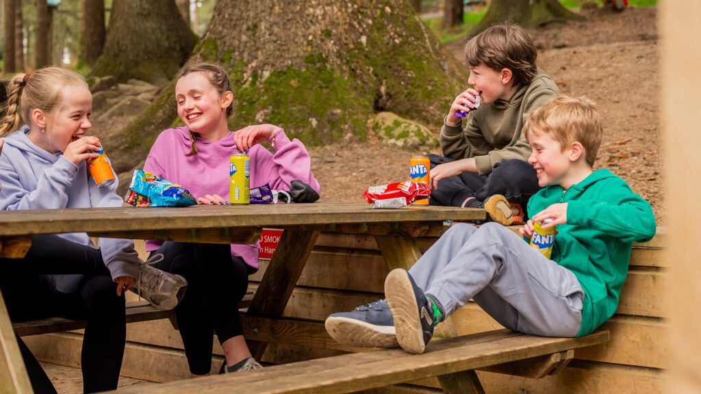Four children sit at a wooden picnic table in a forest, eating snacks and drinking canned sodas, with trees visible in the background.