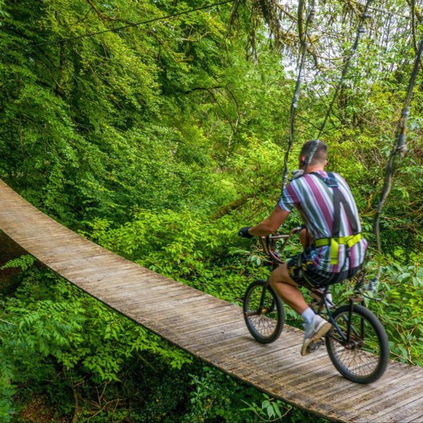 A person enjoys an outdoor escape, riding a bicycle on a narrow wooden suspension bridge surrounded by dense green forest.