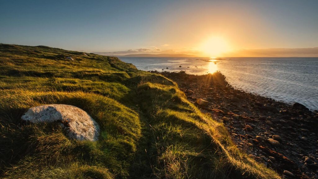 Sunset over a grassy coastline with rocks scattered along the shore and sunlight reflecting on the calm sea, capturing the tranquil beauty of the Summer Solstice Ireland on the longest day of the year.
