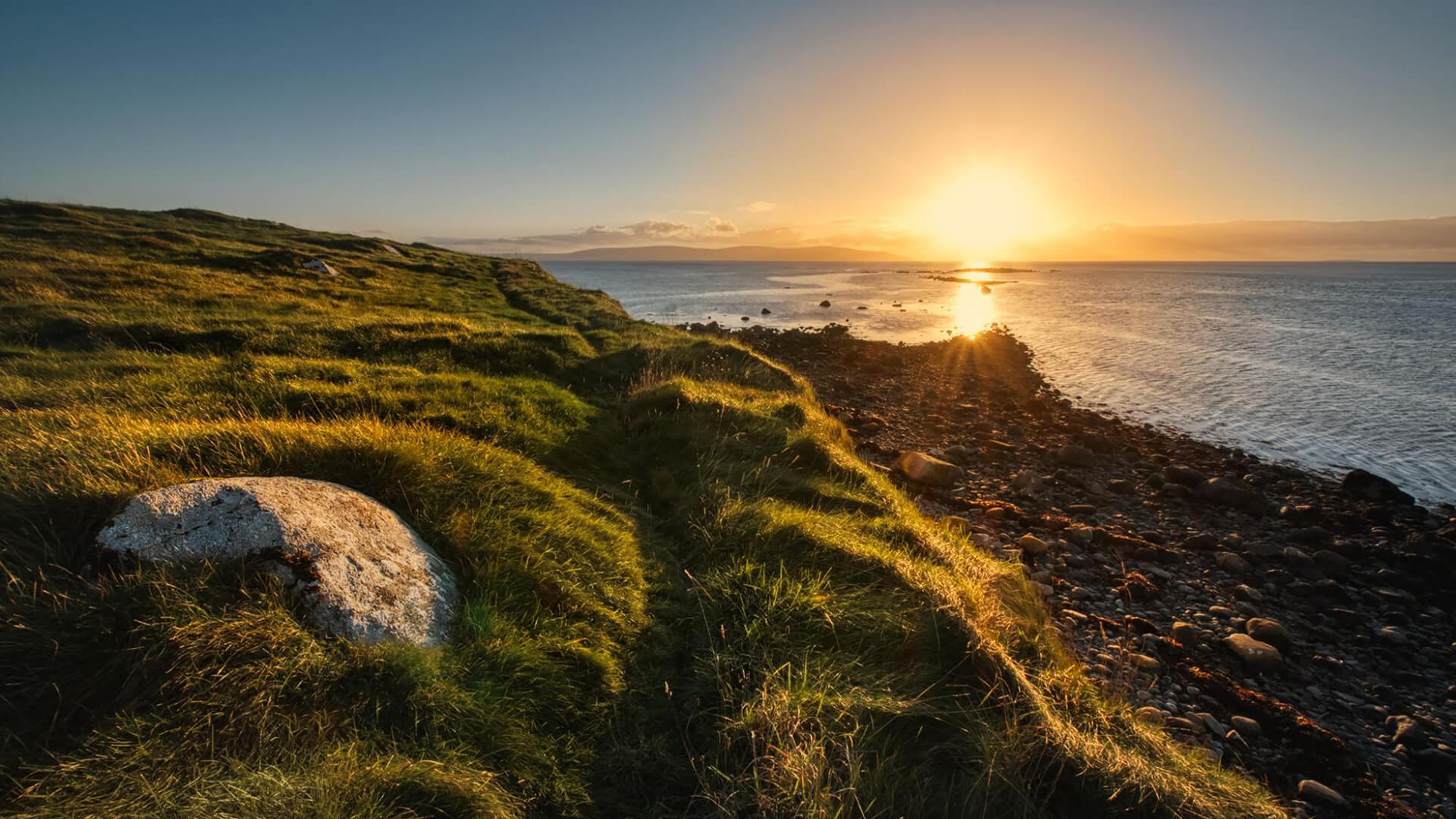 Sunset over a grassy coastline with rocks scattered along the shore and sunlight reflecting on the calm sea, capturing the tranquil beauty of the Summer Solstice Ireland on the longest day of the year.