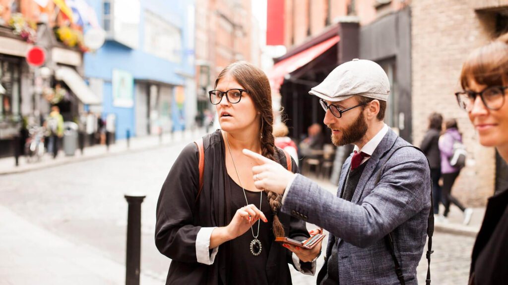 Three people stand on a city street during an Irish summer; one man in a hat points ahead while two women watch attentively. Urban buildings and pedestrians set the scene for lively group activities.
