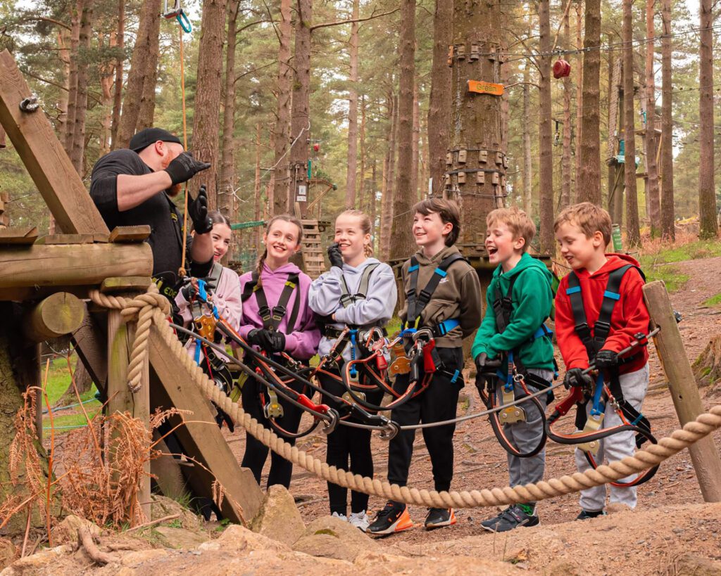 Six children wearing harnesses listen to an instructor at Zipit, an outdoor adventure park surrounded by trees and wooden obstacles—an ideal spot to celebrate World Environment Day with fun and learning in nature.