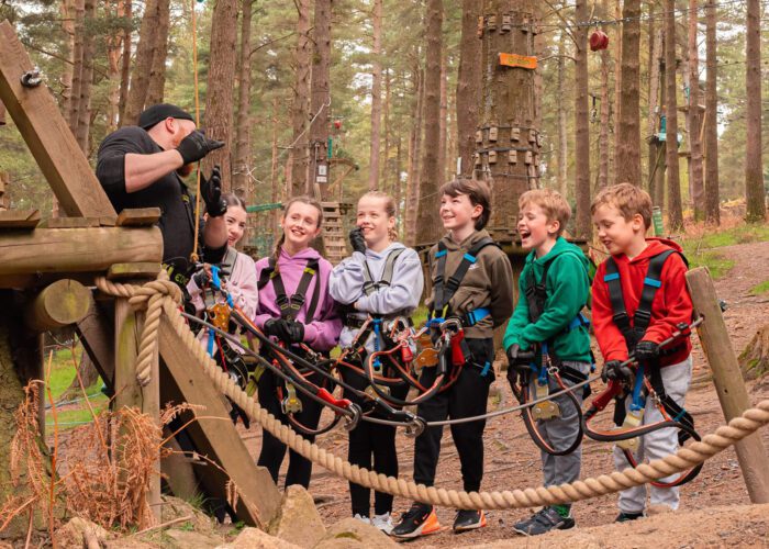Six children wearing harnesses listen to an instructor at Zipit, an outdoor adventure park surrounded by trees and wooden obstacles—an ideal spot to celebrate World Environment Day with fun and learning in nature.