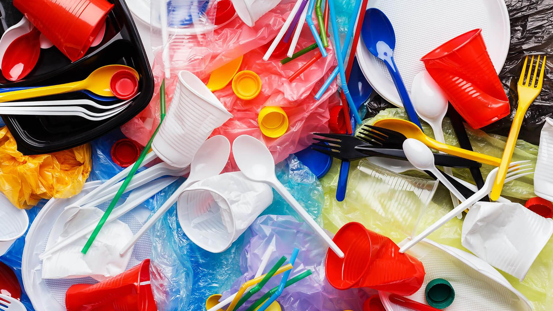 Assorted single-use plastic items, including cups, plates, cutlery, straws, and bottle caps, are scattered on colored plastic bags—highlighting the urgency for change by 2025 and reflecting on the spirit of World Environment Day.