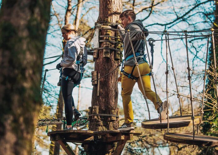 Two people wearing harnesses cross a high ropes course in a forest, walking on suspended wooden platforms attached to trees—an exciting adventure for thrill seekers and a top pick for Hen and Stag Party Ideas in Ireland.