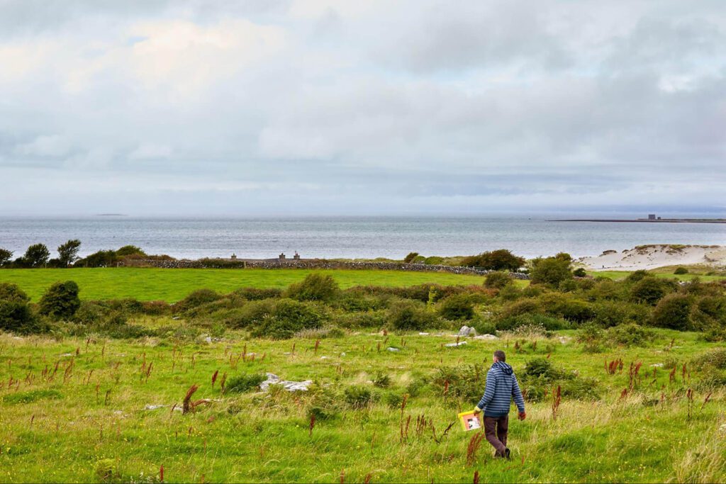A person in a striped jacket walks through a grassy field toward the sea, carrying a small bag—an ideal outdoor escape with cloudy skies above and the distant shoreline stretching beyond.