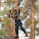 A boy with a safety harness crosses a rope bridge at Zipit adventure park, holding onto a rope above his head—a perfect summer activity for a fun family day out in the forest.