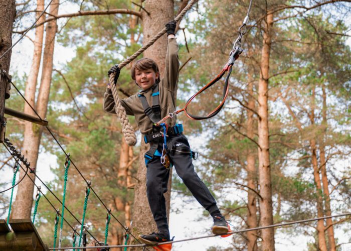 A boy with a safety harness crosses a rope bridge at Zipit adventure park, holding onto a rope above his head—a perfect summer activity for a fun family day out in the forest.