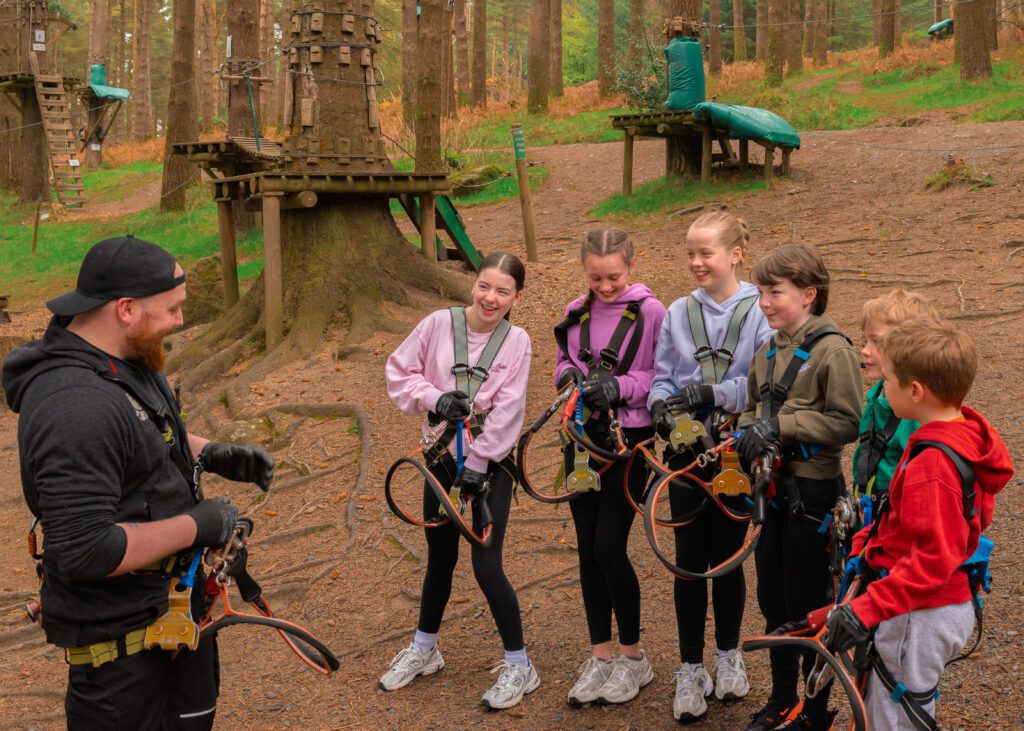 A group of children in harnesses listens to an instructor at Zipit, a perfect summer activity for a family day out, surrounded by trees and climbing equipment in the adventure park.