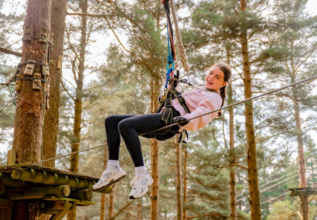 A girl wearing a harness and helmet is riding a Zipit zipline through a forested area, smiling at the camera—a perfect summer activity for a family day out.